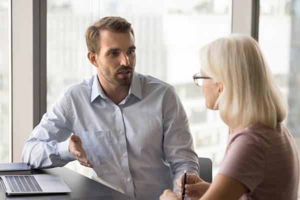 Serious younger manager talking to mature colleague at workplace, discussing collaboration, working on project strategy. Business man meeting with client, partner at office table