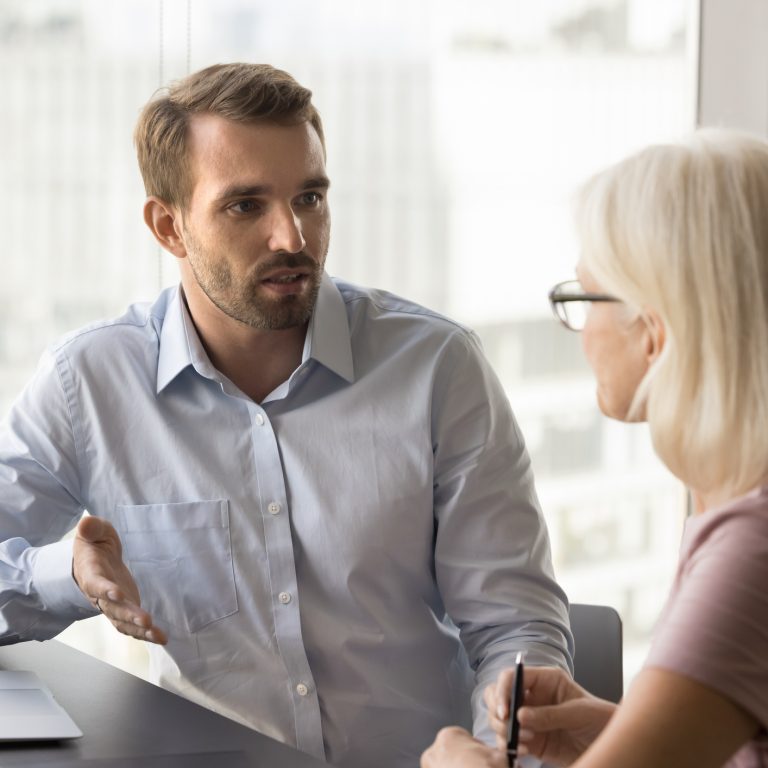 Serious younger manager talking to mature colleague at workplace, discussing collaboration, working on project strategy. Business man meeting with client, partner at office table