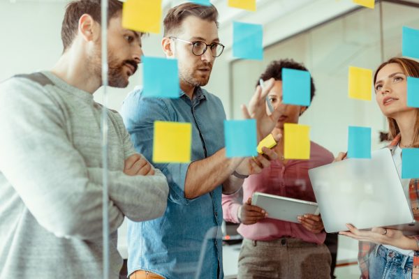 Through glass view of multiethnic male and female executives in casual wear using post it notes while sharing ides at modern office