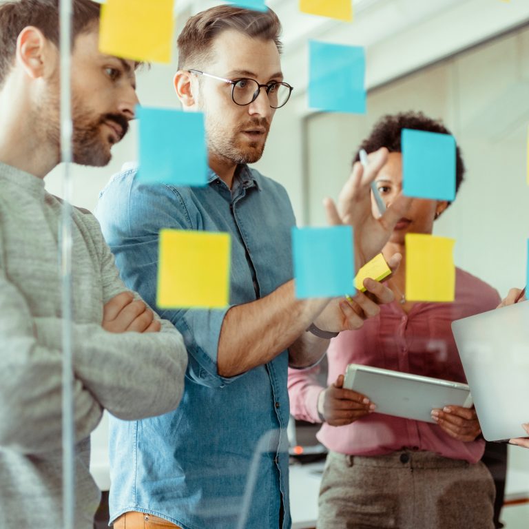 Through glass view of multiethnic male and female executives in casual wear using post it notes while sharing ides at modern office